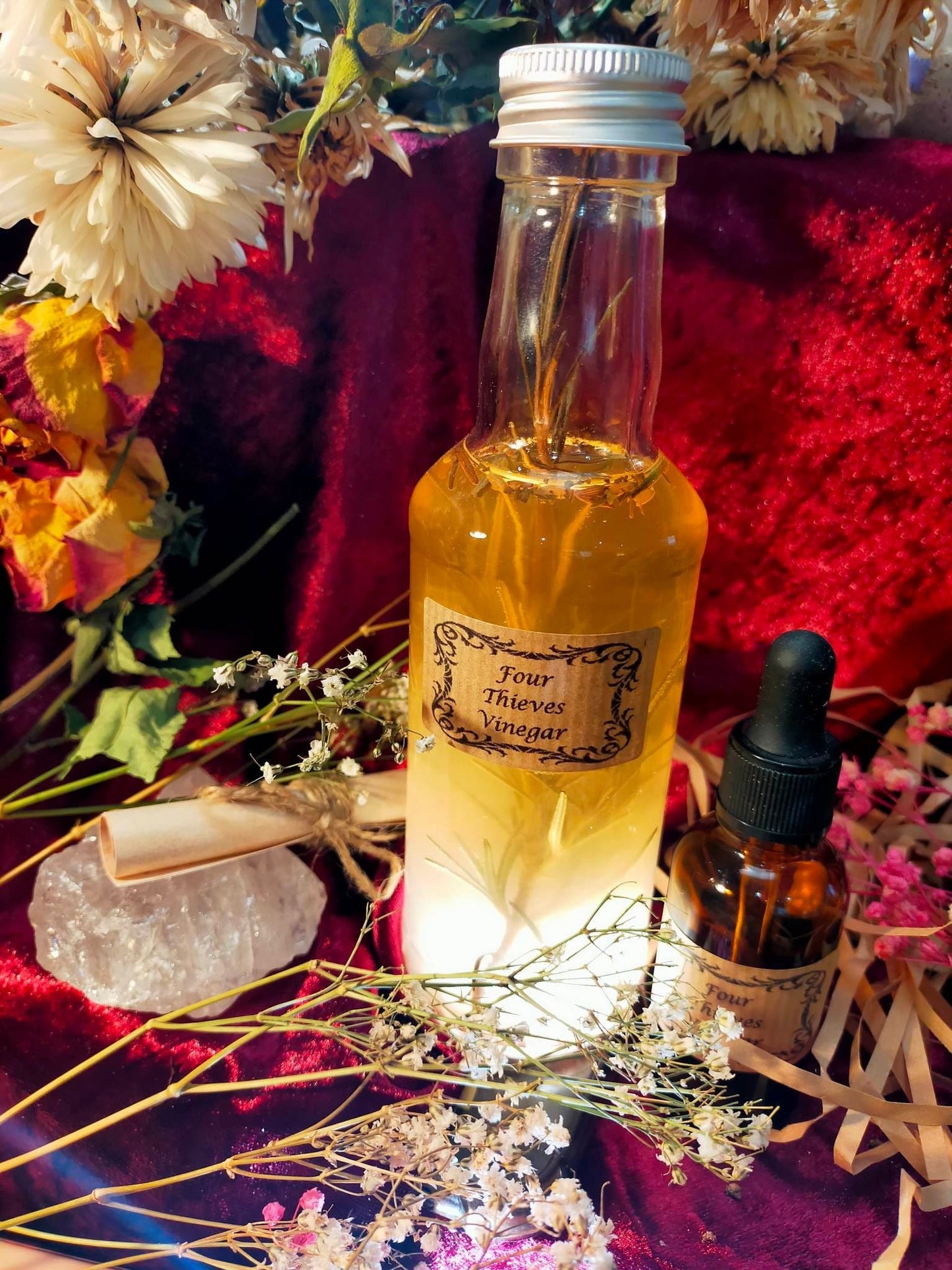 Photo of a bottle of four thieves vinegar with a small vial with dropper labelled 'Four Thieves Vinegar' next to it. There is also a small tied scroll and a large crystal. Dried flowers surround the items.