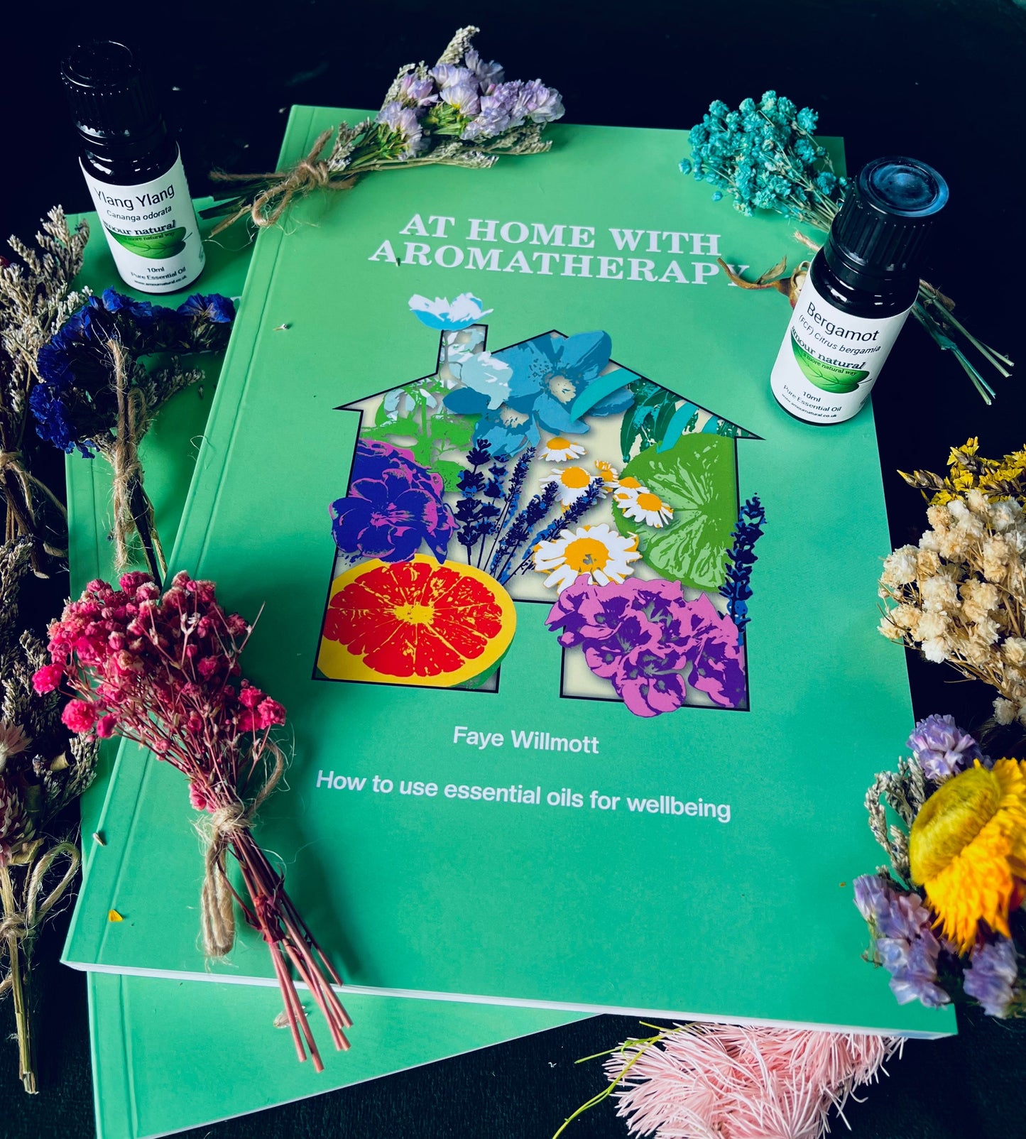 A photo of the book At Home with Aromatherapy, surrounded by herbs, flowers and essential oil bottles.