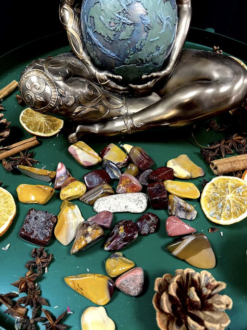 Photo of several mookaite tumblestones amid dried fruit and spices and in front of a Mother Earth figurine.