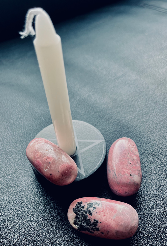 Photo is of rhodonite tumblestones next to a white candle in a small black holder.