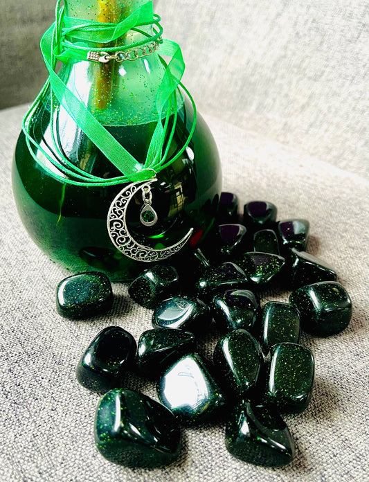 Photo of a number of green goldstone tumblestones in front of an ornate bottle of forest green bath oil.