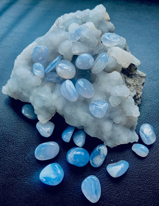 Photo shows a number of small blue chalcedony tumblestones scattered on/around a larger stone.